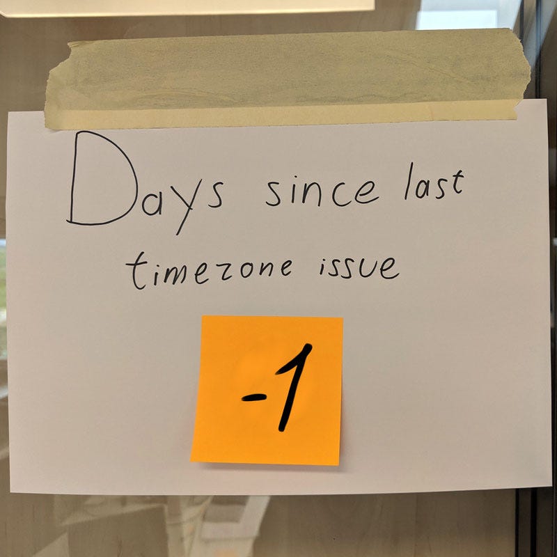 A handwritten sign says “Days since last timezone issue”. A sticky note under that says “-1”.