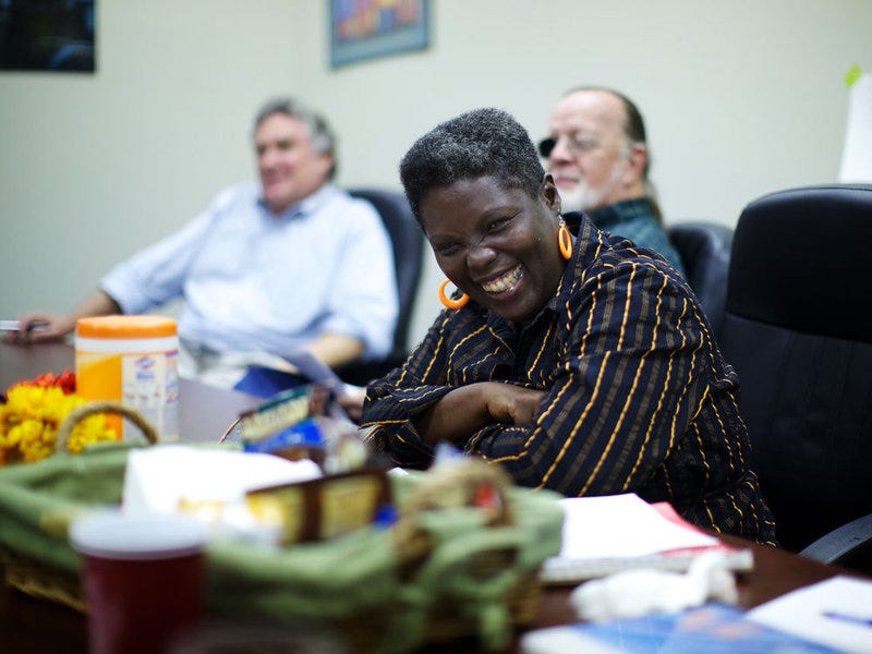 Curtis sits at a cluttered meeting table with two men beside her, smiling with a black, yellow, and brown striped shirt.
