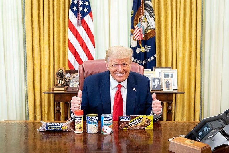 Trump with two thumbs up behind the Resolute Desk with Goya canned goods in front of him.