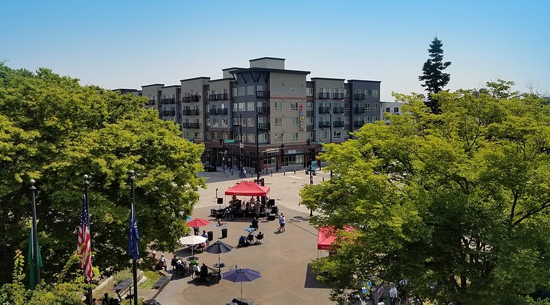 Auburn city plaza under a cloudless sky. Trees and three flag poles are seen in the foreground. Tents and umbrellas are set up with people sitting and mingling. A mixed-use multi-family housing building is visible in the background. 