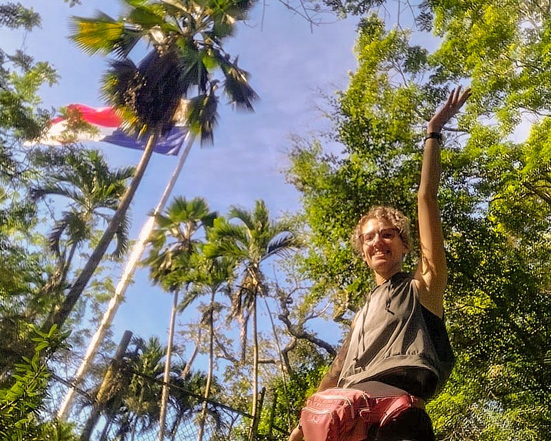 Looking up from the ground the Panama flag can be seen among tall palm trees. A woman in the foreground stands with an arm raised gesturing to it.