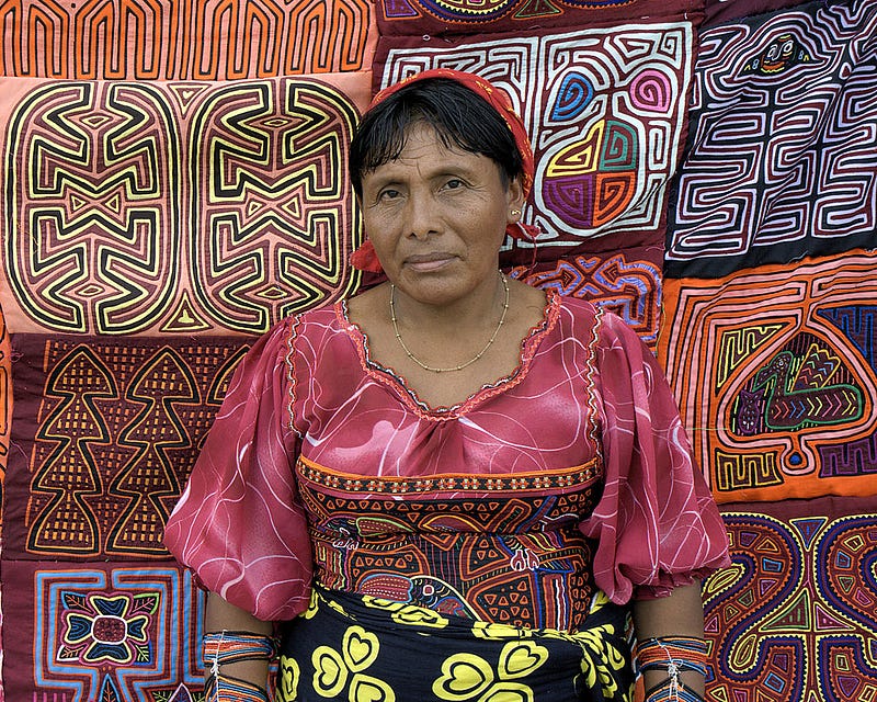 An indigenous woman in a brightly-colored top with an intricate geometric design stands in front of other hand-stitched fabrics with similar colorful designs.