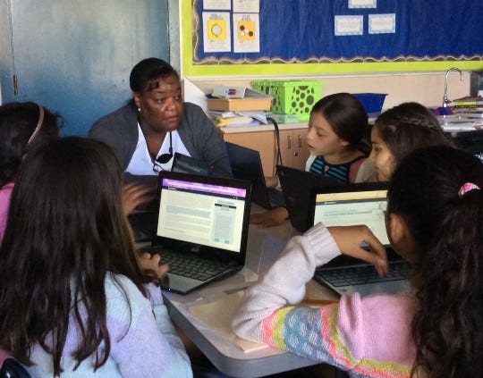 A teacher sits in front of students with their laptops open to a reading lesson. 
