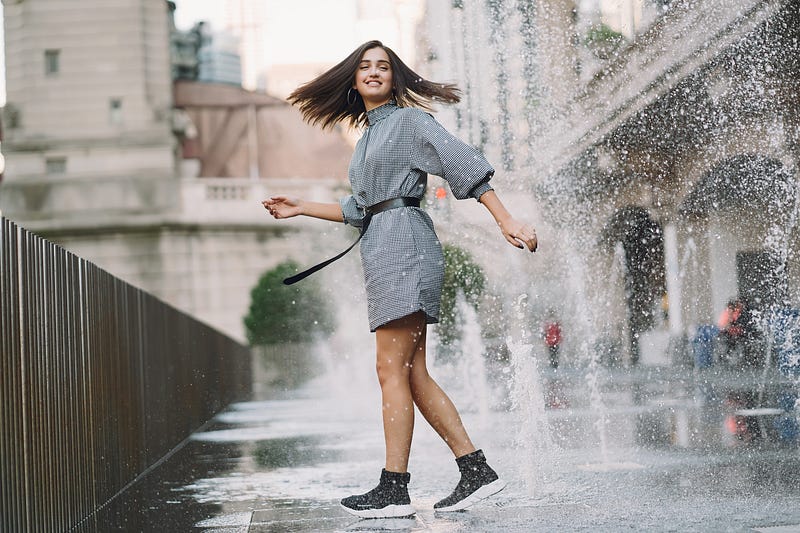 Girl with brown hair and grey dress playing and dancing around on a wet street and having fun.