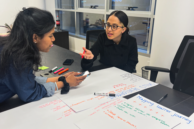 Photo of two students sitting at a table with white poster boards and markers, taking notes and talking with each other.