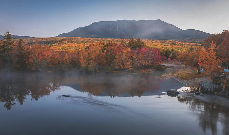 Mount Katahdin in Maine viewed from Abol Bridge with peak reflections on calm water framed by colorful fall foliage.