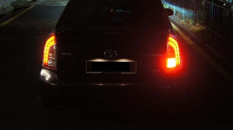 A traseira de um carro, com as lanternas acesas, no escuro de alguma estrada ou viela de condomínio.
