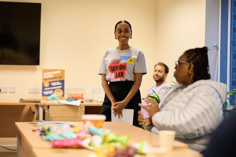 A black woman stands smiling in a branded t-shirt saying Job Design Lab, in front of a table covered in colourful objects. There is a black woman sitting at the table and an Asian man in the background.