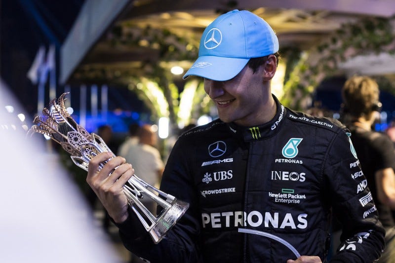 George Russell smiling at the third place trophy he’s holding after he was handed it somewhere in the paddock.