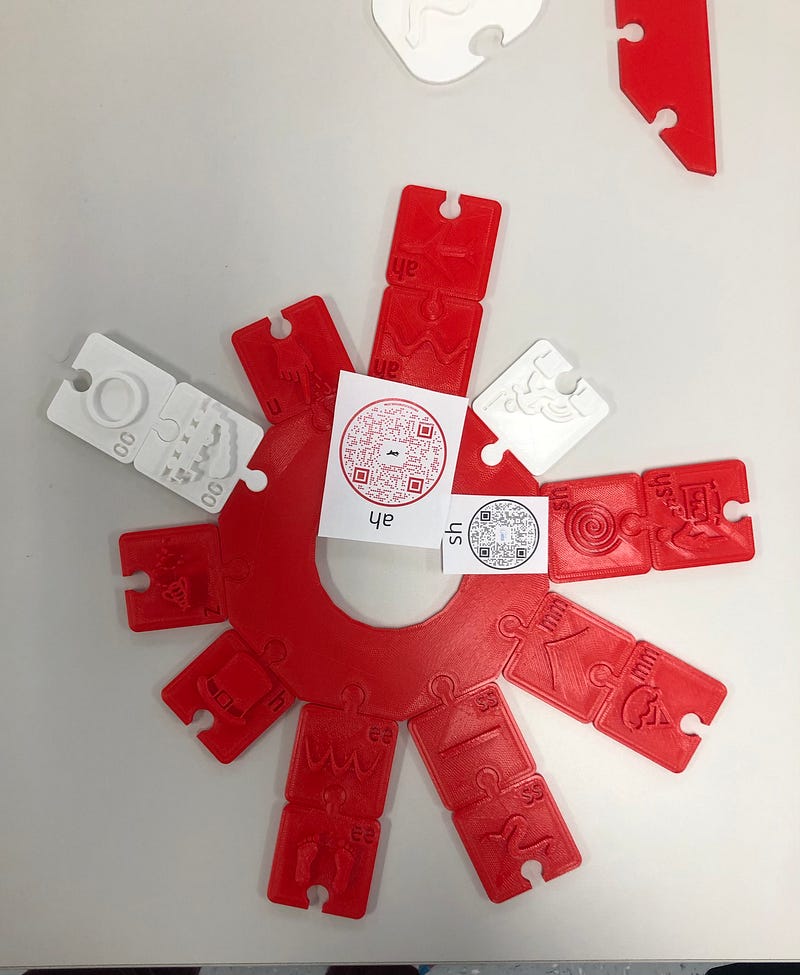 Photo of red plastic 3D-printed puzzle pieces forming a Ling sound puzzle, which is a tactile education tool for young children with hearing or speech difficulties to be able to phonetically pronounce and understand verbal sounds.