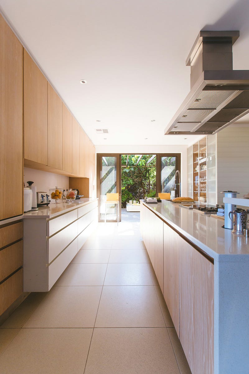 This modern modular kitchen design showcases clean lines, warm wooden cabinetry, and sleek white countertops for a bright and functional space. Natural light flows in through large glass doors, creating a seamless connection between the kitchen and outdoor area. Built-in storage, a streamlined island, and contemporary fittings enhance efficiency and style, making this kitchen ideal for modern homes seeking elegance, practicality, and a refined minimalist interior.