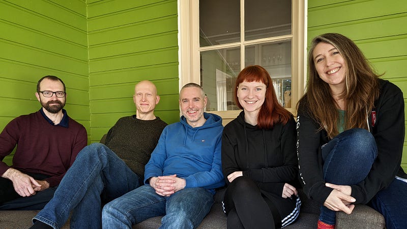 Five people sitting smiling at the camera in front of a window and green painted slats