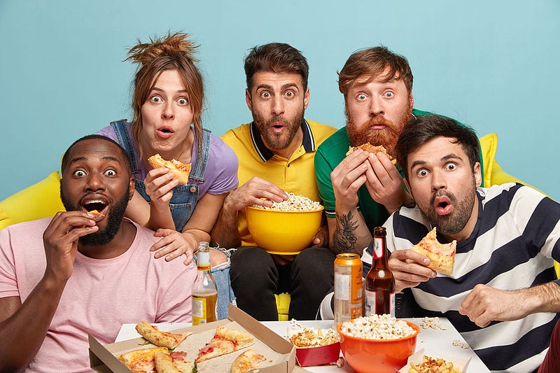 Five funny people eating popcorn and pizza, directly looking at you probable watching video. The screen itself it not visible