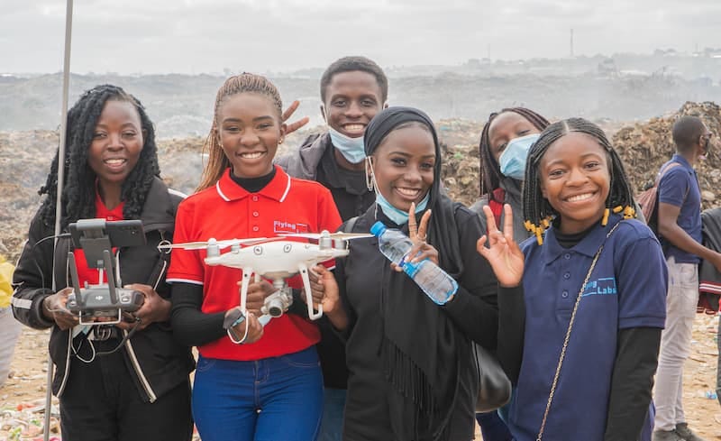 Five girls standing with drones in front of a rocky beach background. Two on the right and a boy behind them are giving peace signs.