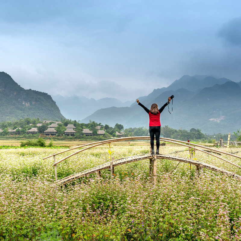A lady standing on a little bridge with a camera in her outstretched arms facing the mountains on the horizon.