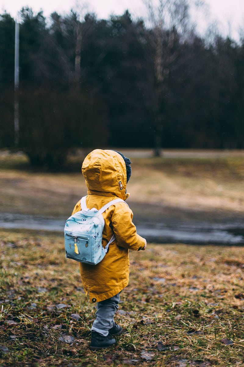 Photo of a child taken from behind. The child is wearing a rucksack and a yellow jacket. He’s outdoors.