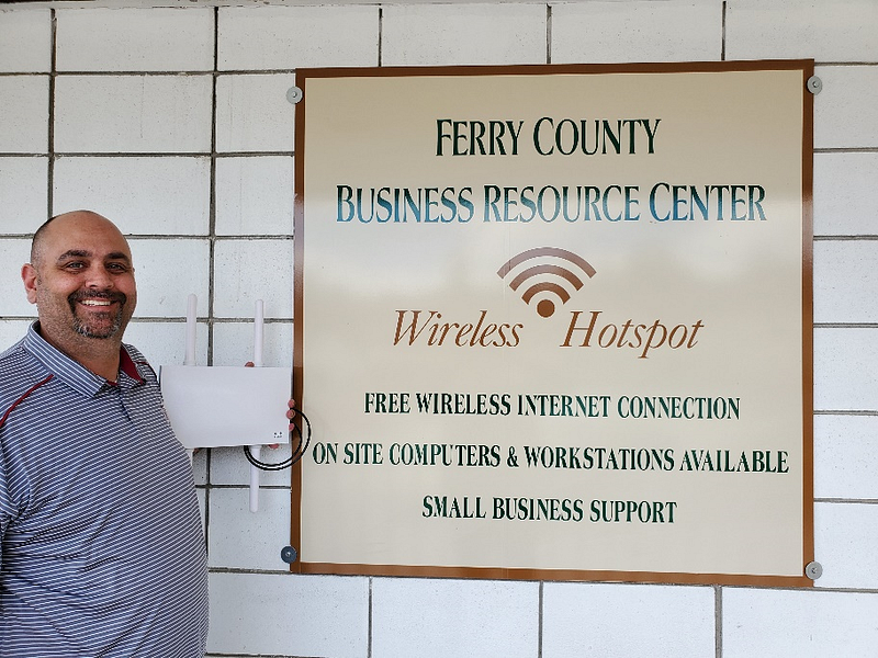 A man stands next to a sign that reads "Ferry County Business Resource Center." The man appears to be holding a piece of connectivity equipment and is wearing a thin striped polo shirt. 