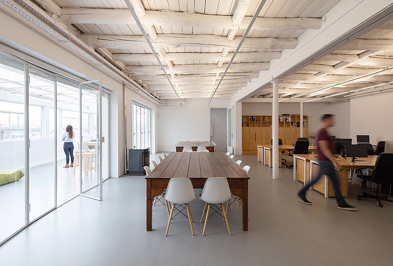 Empty cowork with a big wooden table and white chairs. On the left, a person staring at a window. On the right a person in movement