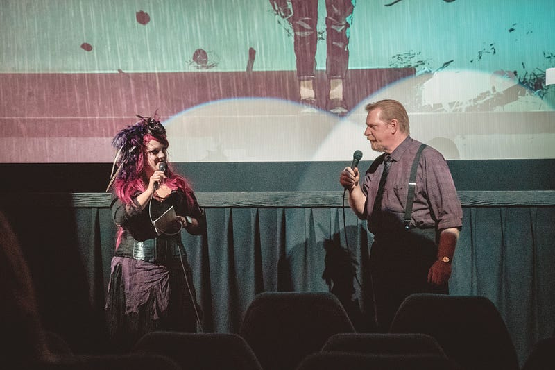 Two people stand on the floor in front of a projection screen. A white woman with dark pink hair is on the left, wearing a feathery headdress and a lace-and-corset outfit. She holds a microphone a notes while smiling at the white man wearing suspenders on the right.