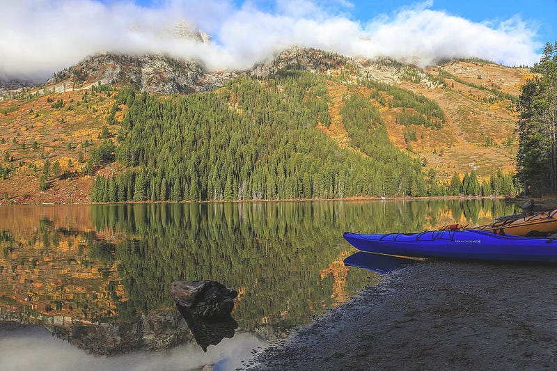Colorful kayaks rest along the shore of String Lake in Grand Teton National Park during autumn, surrounded by golden trees and mountain views — photograph by Dan Sproul.