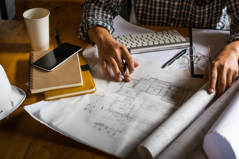 This image captures an architect reviewing detailed floor plans at a well-organized workspace. Rolled blueprints, notebooks, a keyboard, and essential tools highlight the precision and focus required in architectural planning. The scene reflects creativity, technical expertise, and hands-on design development — perfect for showcasing professional architecture, construction planning, and project drafting processes.