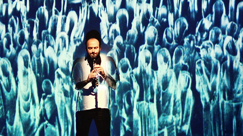 A man plays clarinet in front of a screen with ghostly illustrations of people.