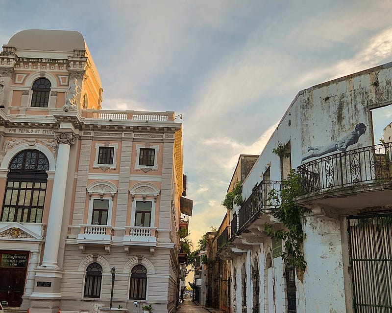 A well preserved white and pale pink colonial style building sits next to a dilapidated building. All that remains are weathered white walls with grafiti and iron teraces.