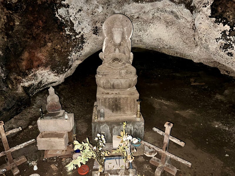 Statue with flowers in front and a smaller statue to the side in front of mouth to another cavern.