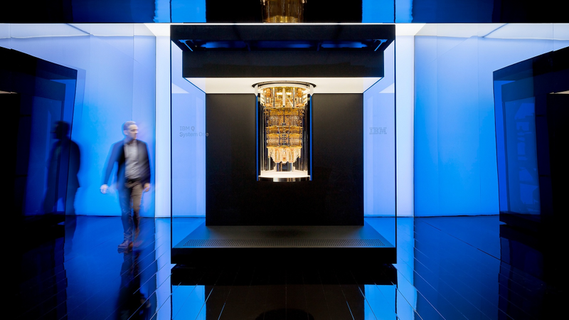 A room with blue lighting. At the centre of the room, a large, bronze piece of intricate tech hangs from the ceiling. A person walks around the centrepiece, but is blurry in the image.