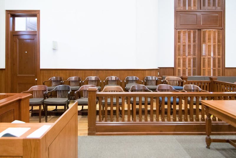 A photo shows a jury box in an empty courtroom. Fourteen wooden chairs are lined up in two rows. Shuttered windows are shown to the right behind the jury box and a door is shown to the back left. A wooden banister partially surrounds the jury box.