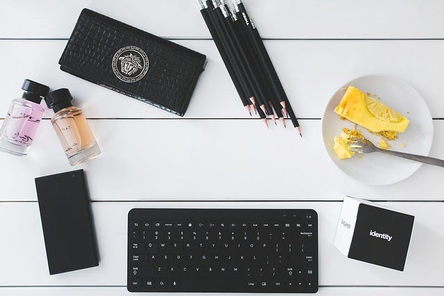 A white table with a Versace wallet, a bunch of black pencils, a black keyboard, a phone and two travel size perfums.