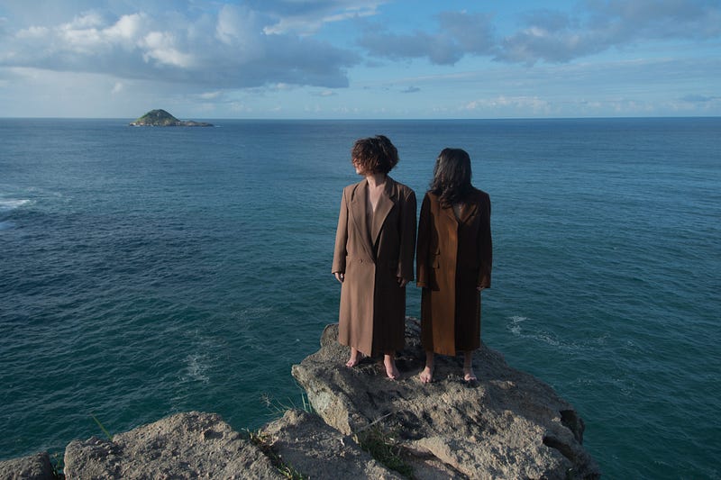 duas mulheres com roupas na cor marrom, de costas para o mar olhando para o lado esquerdo