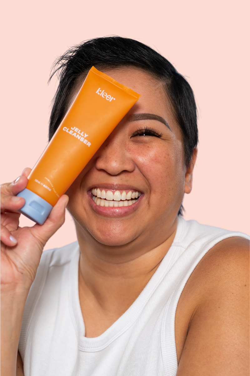 An image shows an Asian woman with flawless skin smiling brightly as she holds a tube of Kleer skincare jelly cleanser in her hand. She exudes confidence and radiance, with her short dark hair. The focus is on the product in her hand, which is a bright orange tube with perwinkle lid filled with a clear gel-like substance, indicating its refreshing and gentle nature.