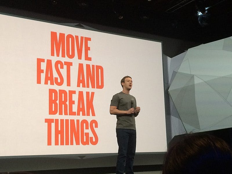 Mark Zuckerberg on stage holding a microphone, standing in front of a screen that says “move fast and break things”