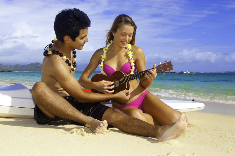 Girl in pink bikini playing the ukulele. Boy is helping her. They are sitting at the beach.