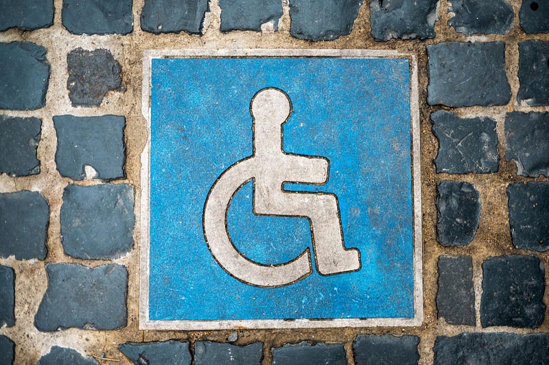 Accessibility symbol painted on pavement, showing a wheelchair icon embedded in a blue square among stone tiles.