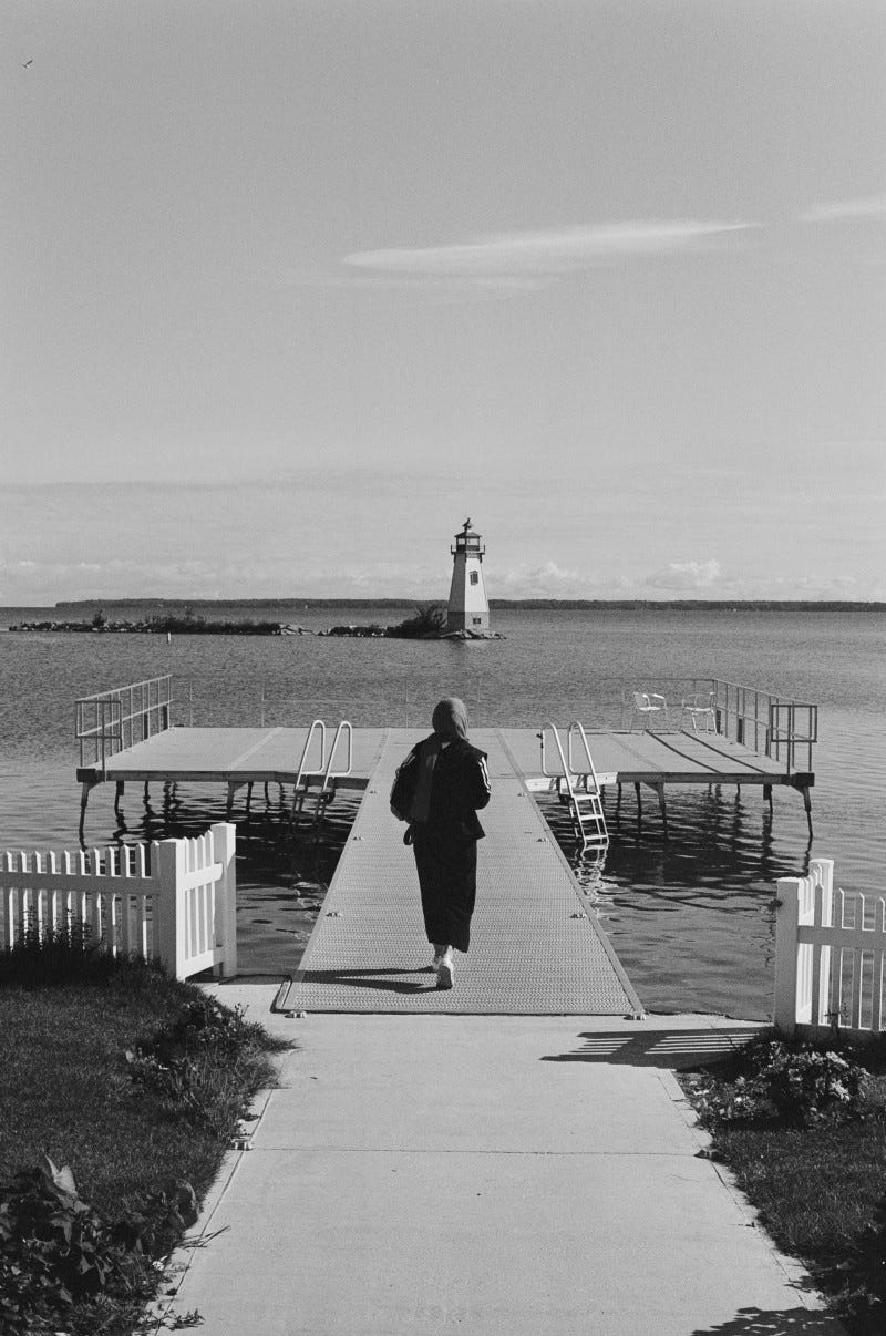 A film photo of a woman in scarf walking towards a pier. We can see a lighthouse in the distance.