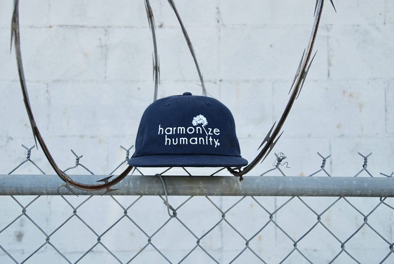 Picture of a baseball cap on top of a chainlink fence. The hat is blue with white writing that reads “harmonize humanity”. There is some barbed wire behind it.