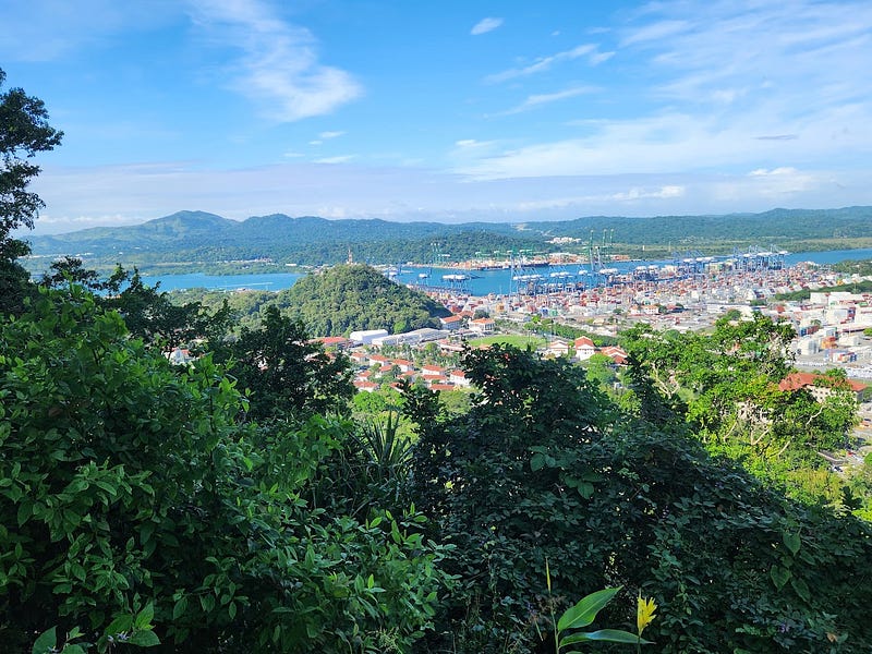 Landscape view from a mountain top. In the foreground are green treetops, above them a shipyard filled with shipping containers, further out the Panama Canal, blue and dotted with ships sits in front of a green mountain range.
