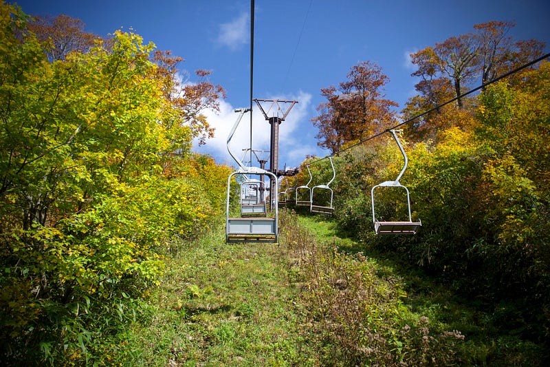 The bright yellow trees, green grass, and blue sky contrast against the grey and rusted Mt. Gassan Ski Lift on the way up Mt. Ubagatake