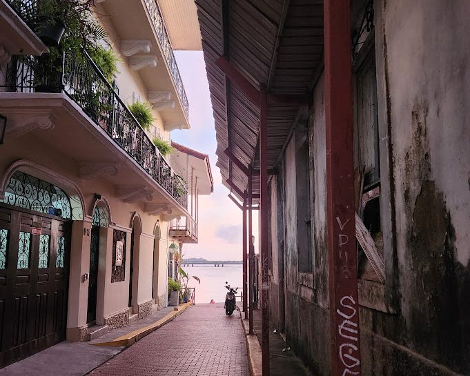 A narrow brick alleyway running between two buildings. One nicely kept and the other worn and graffitied. At then end sits a motorcycle and beyond that the ocean.