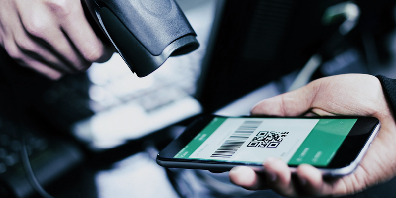 A hand holds a scanner pointing towards a phone being held by another person. On the phone is a barcode and a QR code.