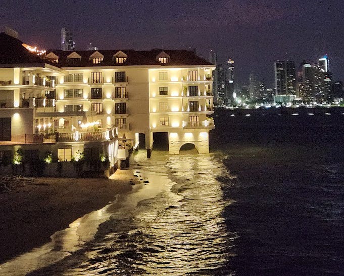 A large white, lighted hotel juts into the ocean, the lights reflected in the wave on the shore.