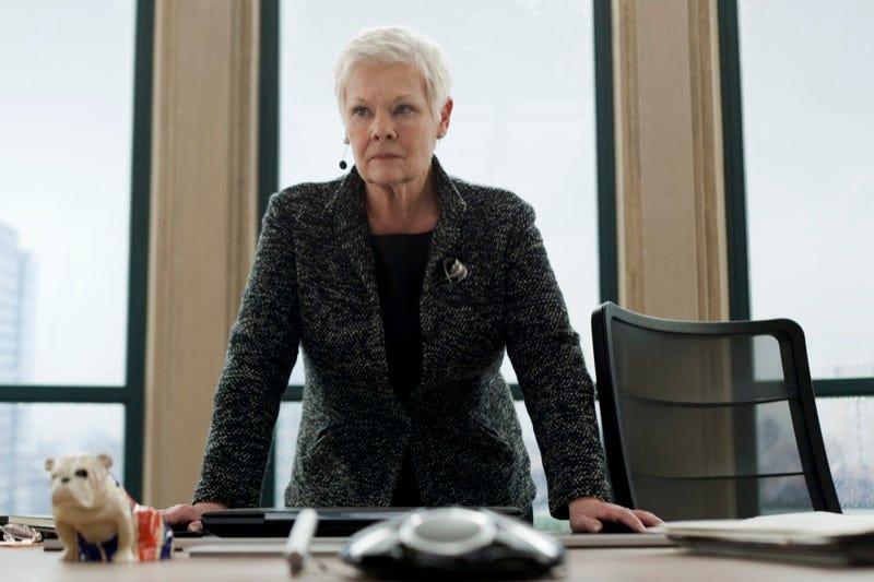 An elderly woman , Judi Dench stands leaning over her desk slightly with a determined looking expression on her face. She has a microphone attached by her ear and is clearly directing someone.