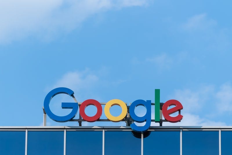The colorful Google logo atop an office building, set against a cloudy blue sky.