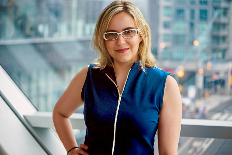 Sarah Kurchak, a white woman with blonde hair and white glasses, smiles into the camera for a portrait. She wears a dark blue shoulderless zip-up top and stands in front of a window that looks out over a city junction.