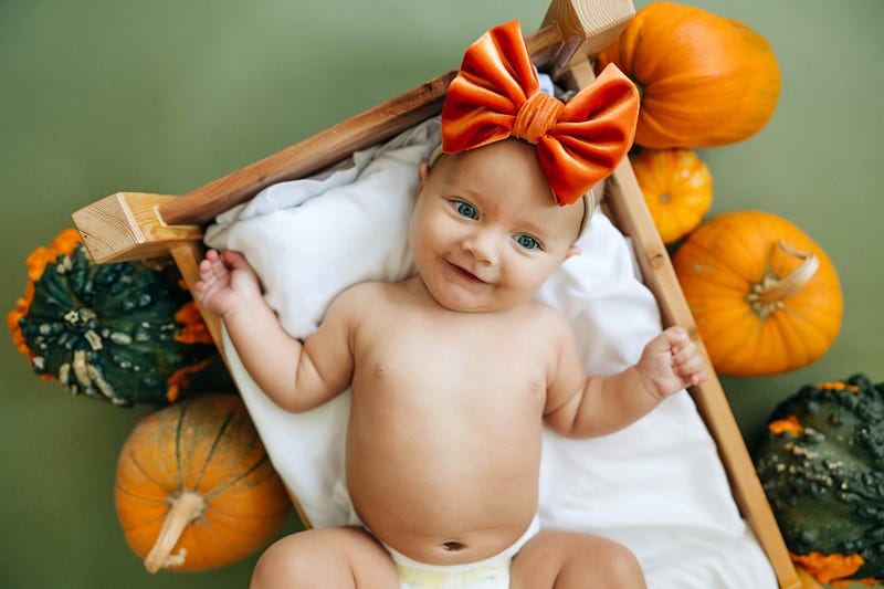 Baby with a diaper and big orange bow, lying on a basket surrounded by pumpkins.