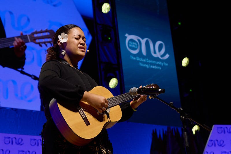 A woman with a white flower in her hair sings while playing the guitar on a stage.