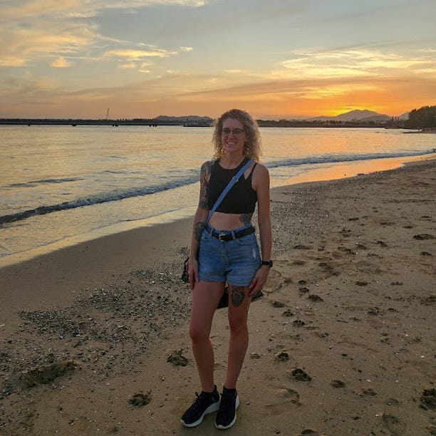 The author, a young woman, stands on a beach at sunset in front of an orange hued sky as the sun sinks behinds a silhouetted mountain.