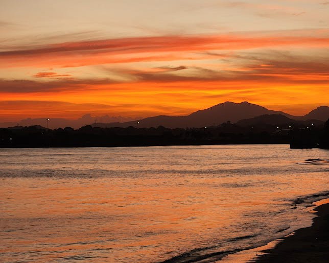 A bright orange and yellow sunset sky over a silhouetted mountain range and reflected in the ocean.